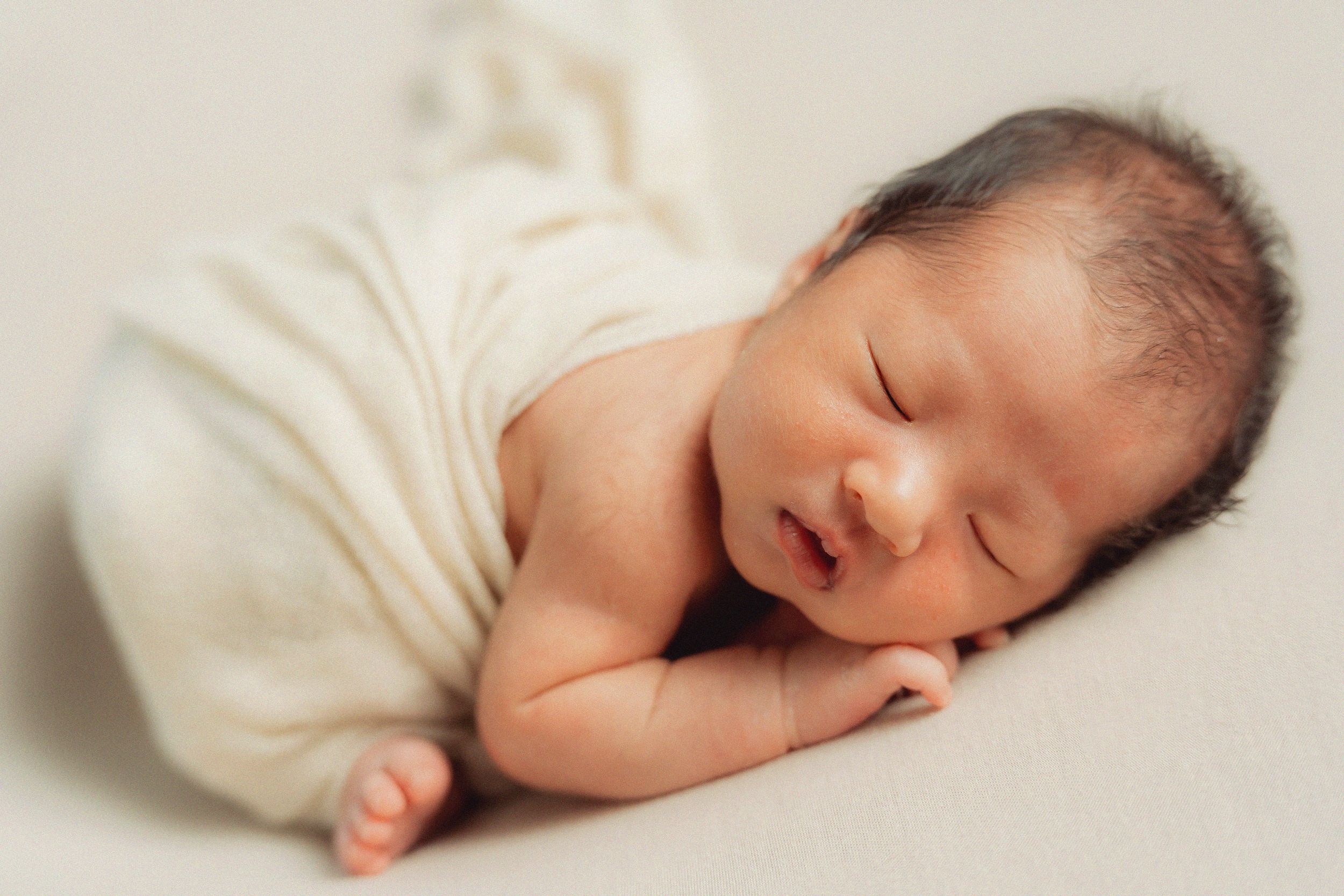 Peaceful sleeping newborn baby wrapped in soft white fabric against cream background.