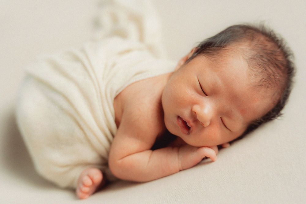 Peaceful sleeping newborn baby wrapped in soft white fabric against cream background.