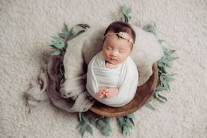 Newborn baby sleeping peacefully in wooden bowl with green foliage accents.