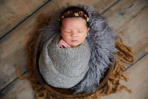 A sleeping newborn wrapped in gray swaddle with a floral headband on wooden floor.