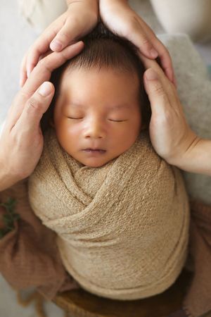 Overhead view of sleeping baby wrapped in beige fabric with gentle hands framing the shot.