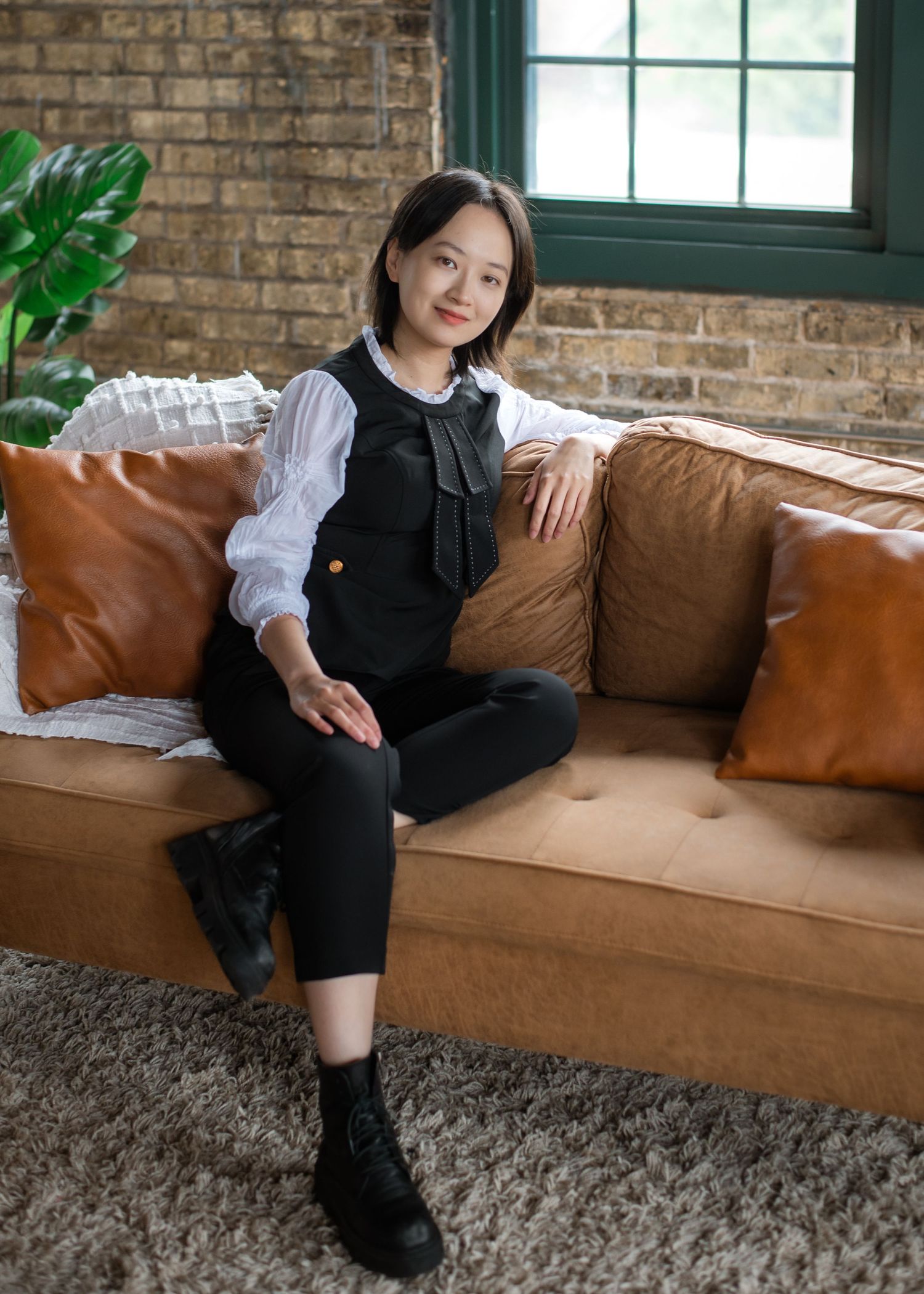 Casual outfit with black overalls and white blouse on a brown leather couch against exposed brick wall and green window.