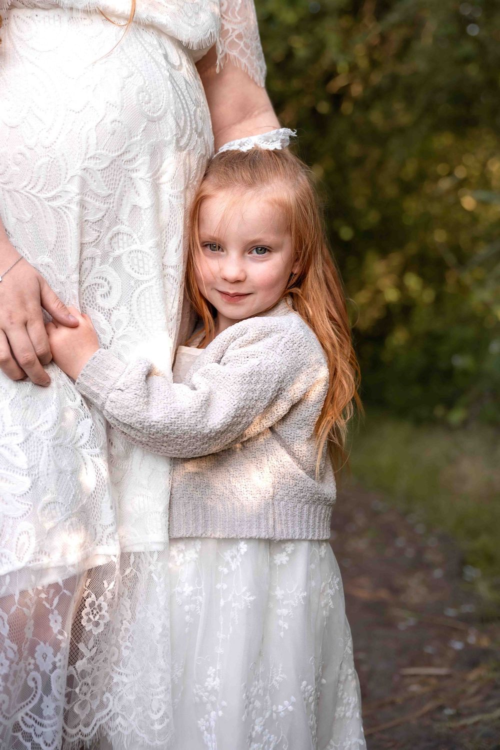 A child in a delicate white lace dress stands outdoors against a natural background.