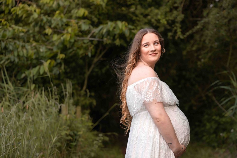 A bride in a lace white dress poses gracefully in a natural outdoor setting with greenery in the background.