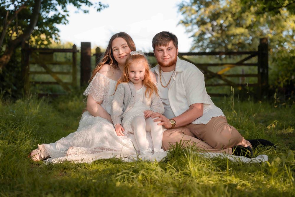 A family wearing white sits together on grassy ground during a golden hour outdoor photo session.