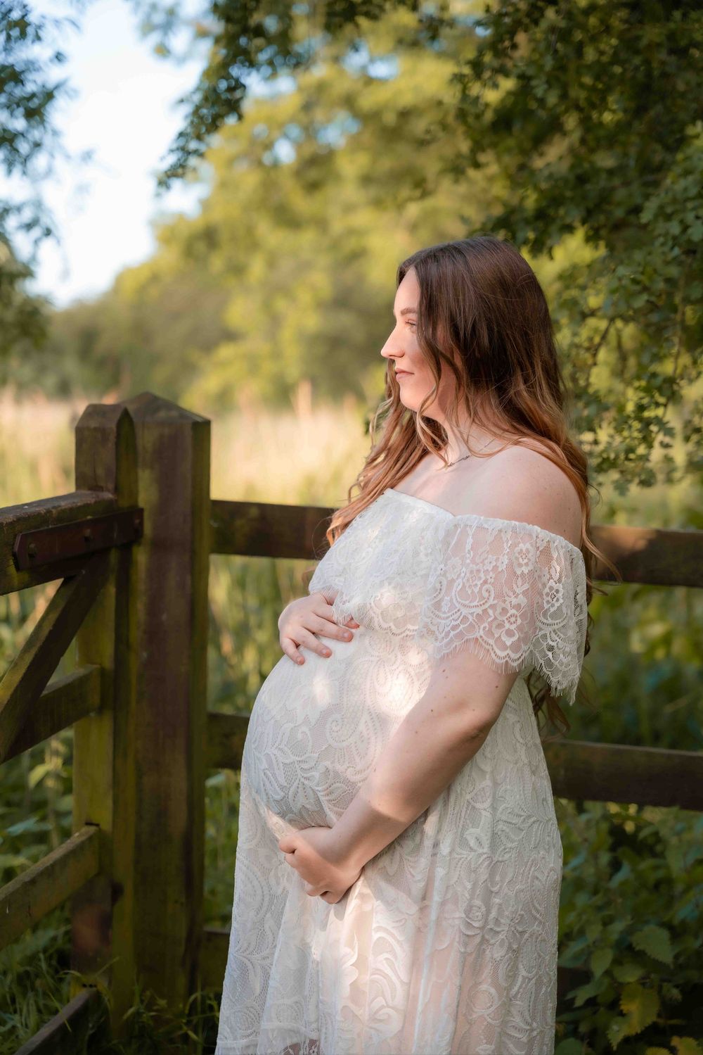 A figure in a flowing white lace dress stands near a rustic wooden fence in a sunlit garden setting.
