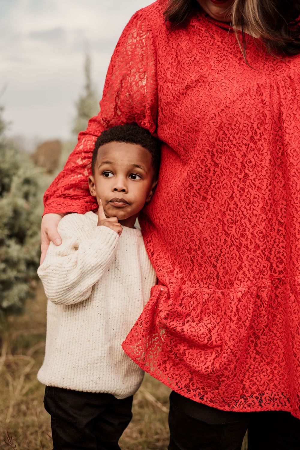 A young child in a cream sweater stands close to someone wearing a red lace dress in an outdoor setting.