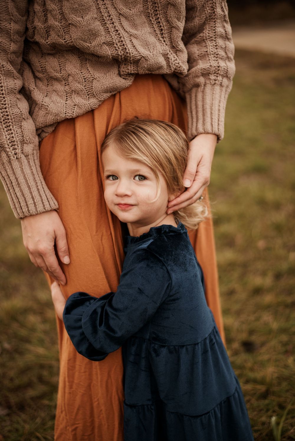 A small child in a navy dress hugs close to someone wearing a brown sweater and orange skirt outdoors.