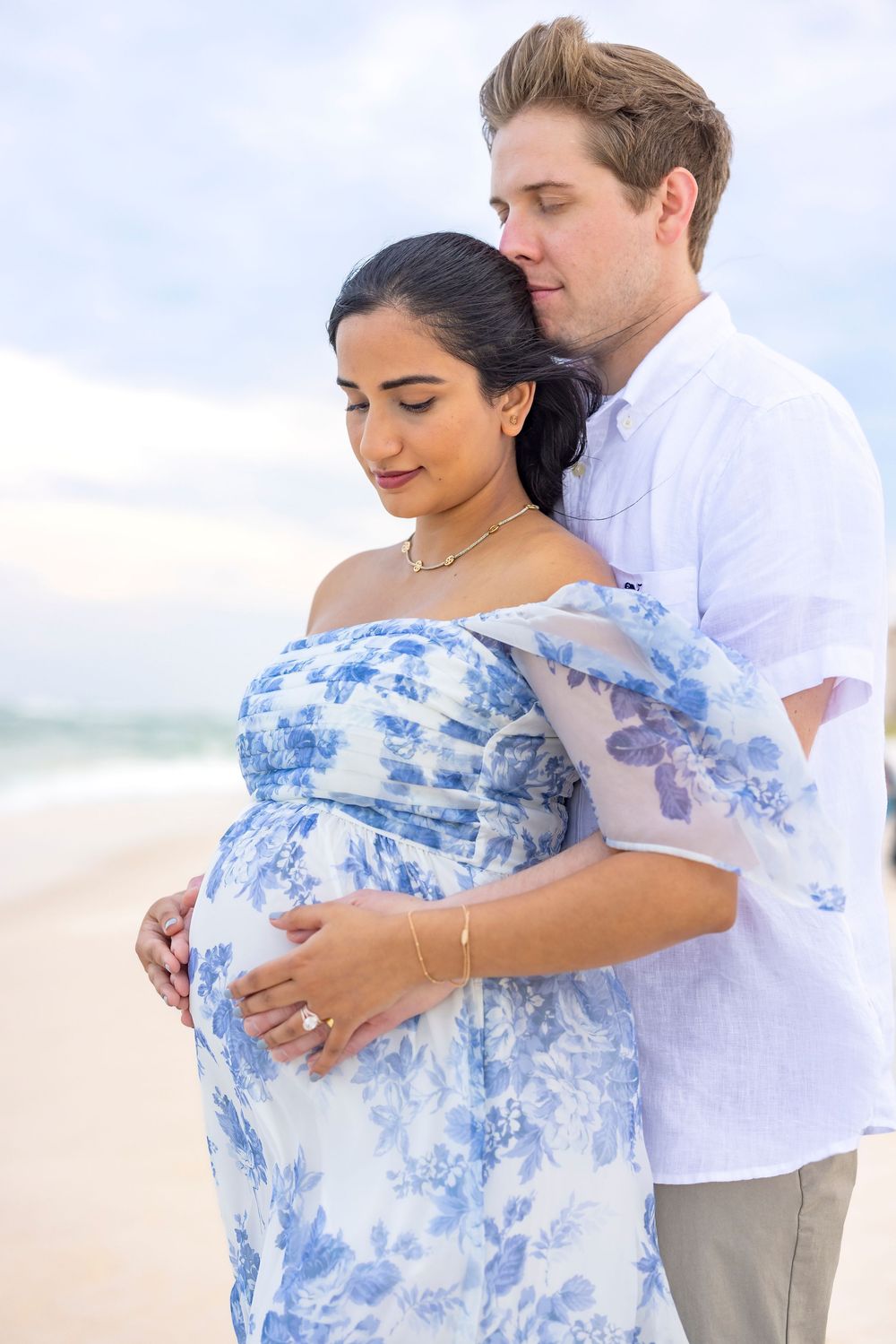 A couple embraces on a sunny beach while wearing white and blue flowy clothing during a romantic photoshoot.