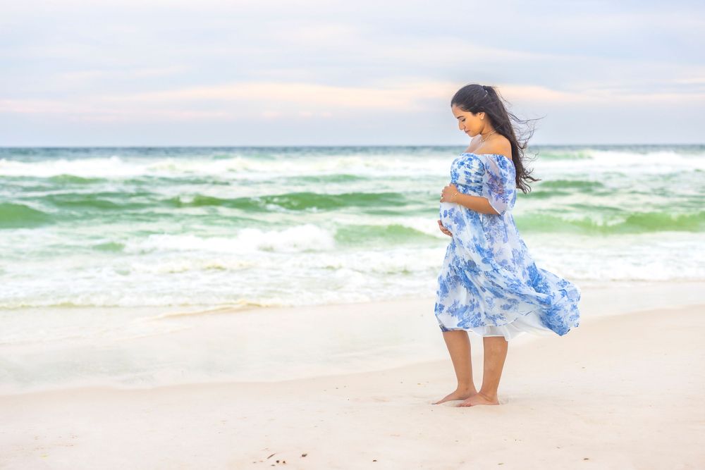 Woman in blue floral dress walks along white sandy beach at sunset with waves crashing in background.