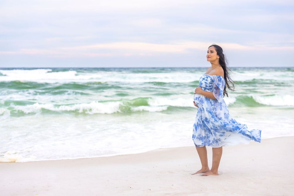 A woman in a flowing blue and white dress walks along the shoreline of a beach as waves crash nearby.