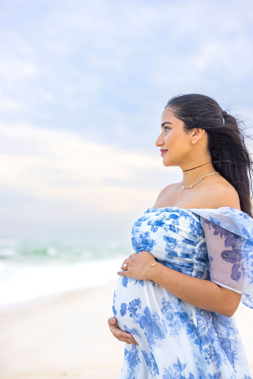 A woman in a white and blue tie-dye dress smiles while standing on a beach with wind blowing through her hair.