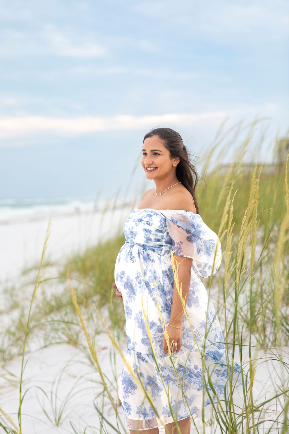 A person in a blue and white floral off-shoulder dress poses among beach grass and sand dunes on a cloudy day.