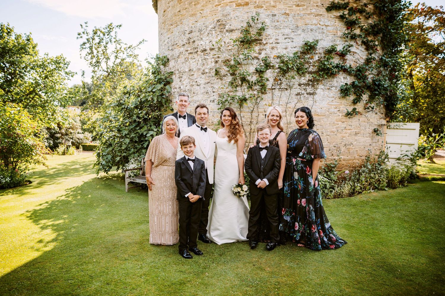 Wedding party members gather outdoors on a sunny day near a rustic stone wall and green lawn.