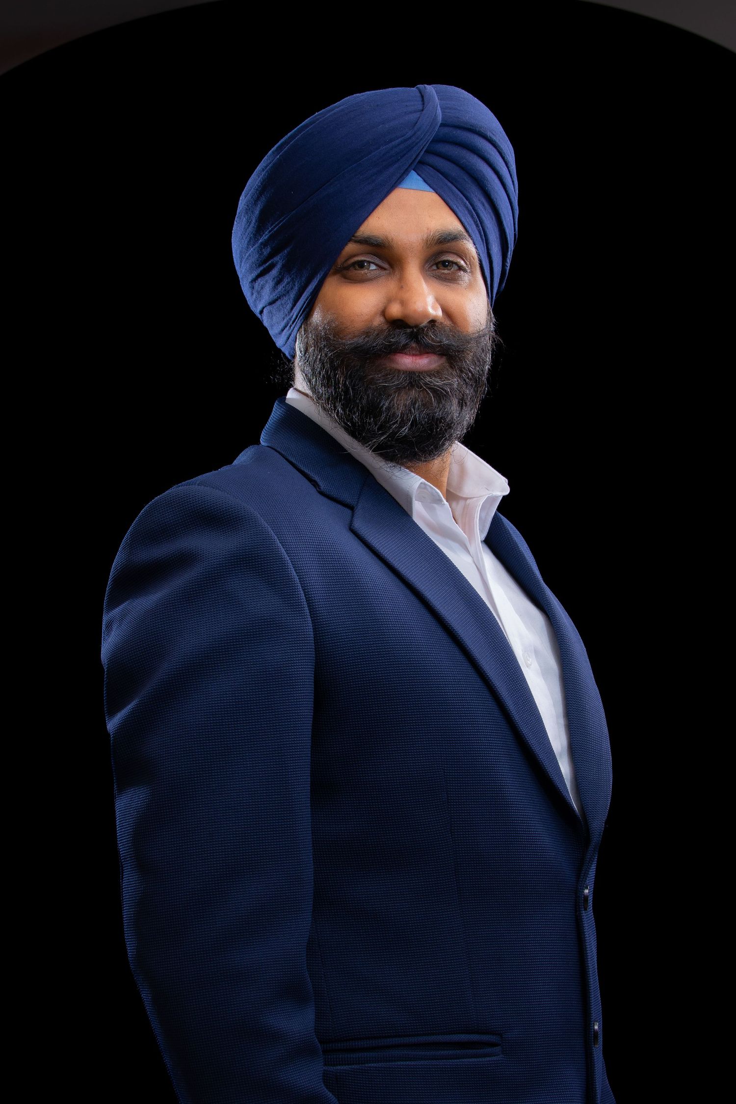Professional headshot portrait of a Ishdeep Singh wearing a navy suit and blue turban against a black background.