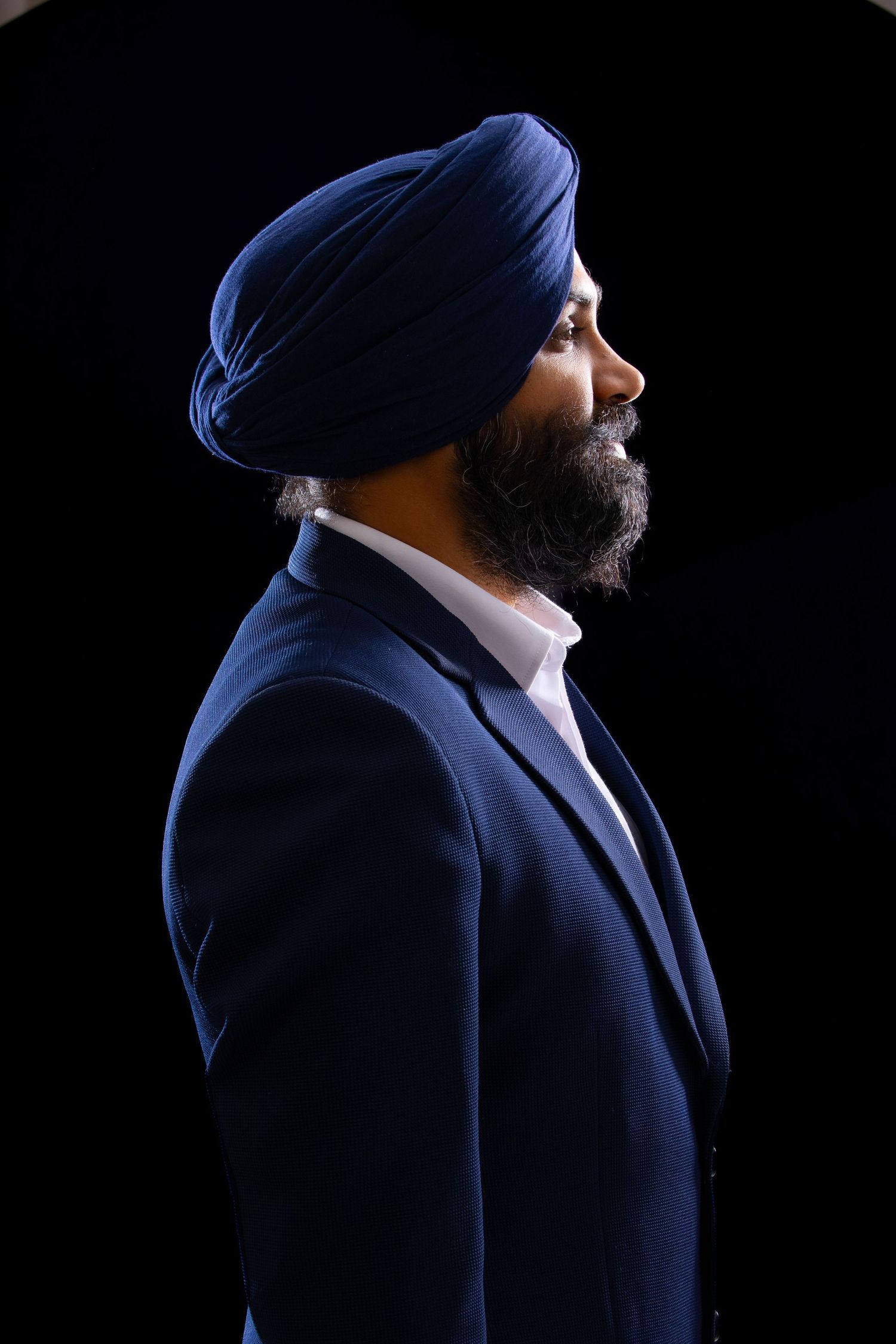 Profile view of Ishdeep Singh wearing navy blue turban and suit against dark background in dramatic lighting.