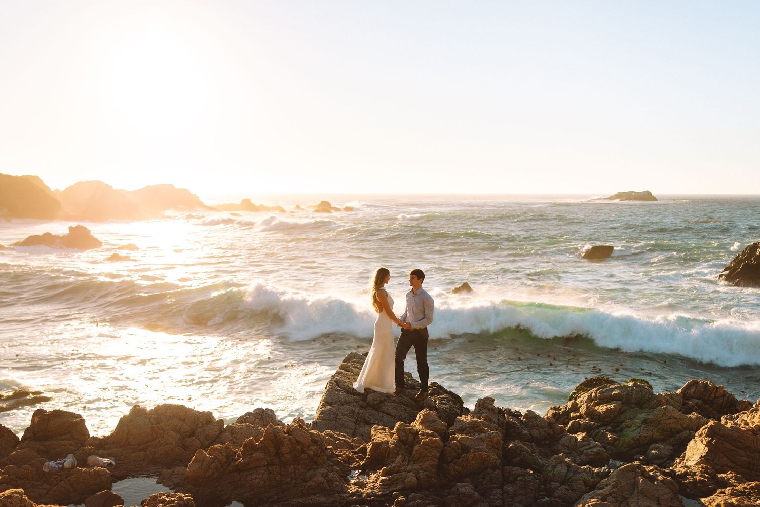 Silhouetted figures stand on rocky coastline during golden sunset as waves crash against California shore.