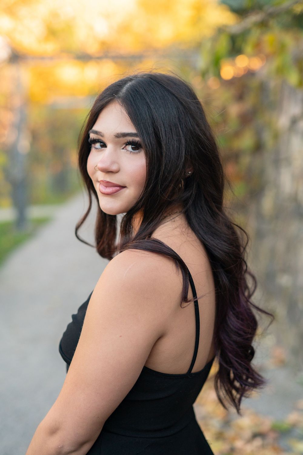 A woman in a black top poses outdoors during autumn with golden foliage in the background.