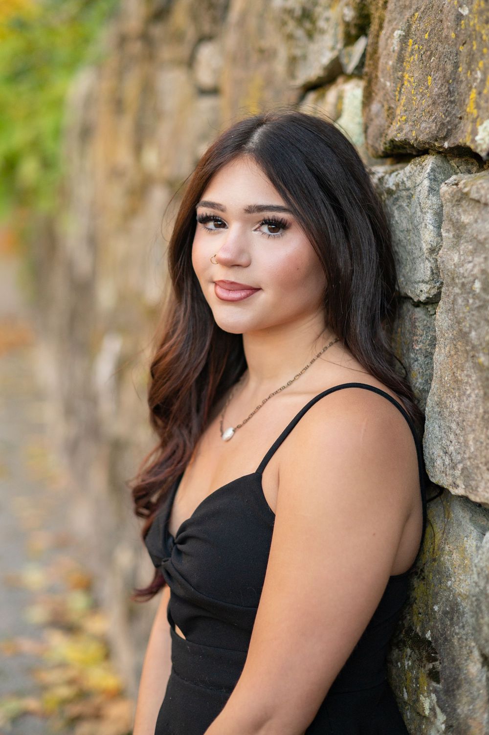 Portrait of someone in a black dress posing against a stone wall outdoors with natural lighting and greenery in background.