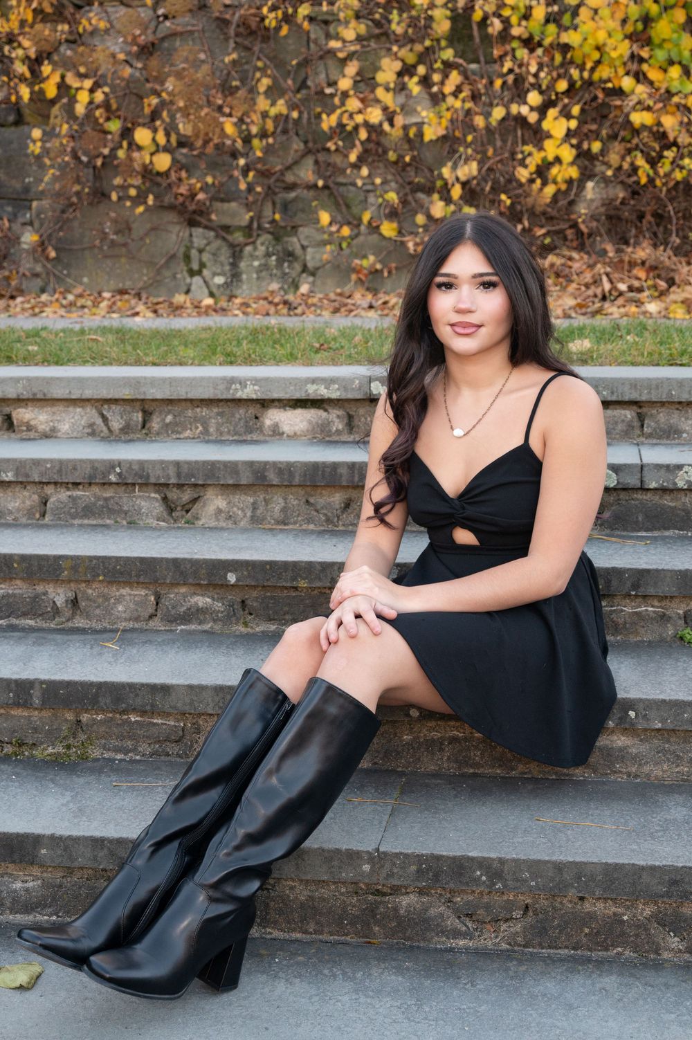 Stylish black dress and boots pose on concrete steps with autumn leaves in background.