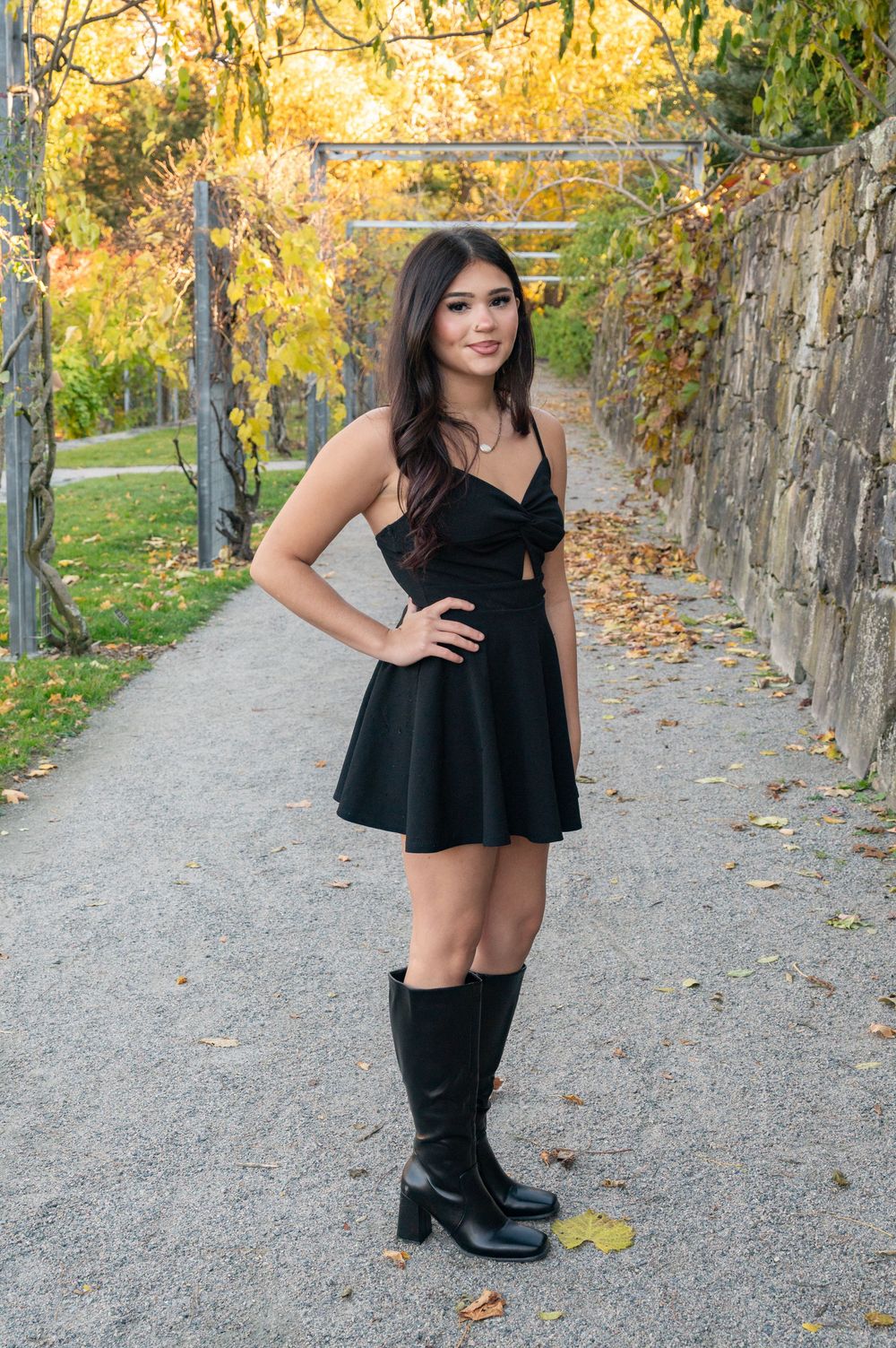 A person in a black dress and knee-high boots poses on a path surrounded by autumn foliage and stone walls.