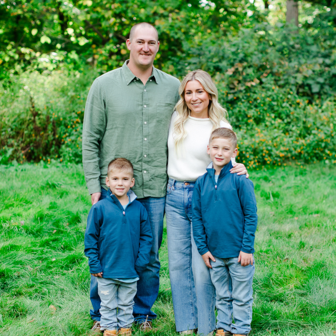 Family of four posing outdoors on green grass with trees, parents and two young boys in a Family Mini Session