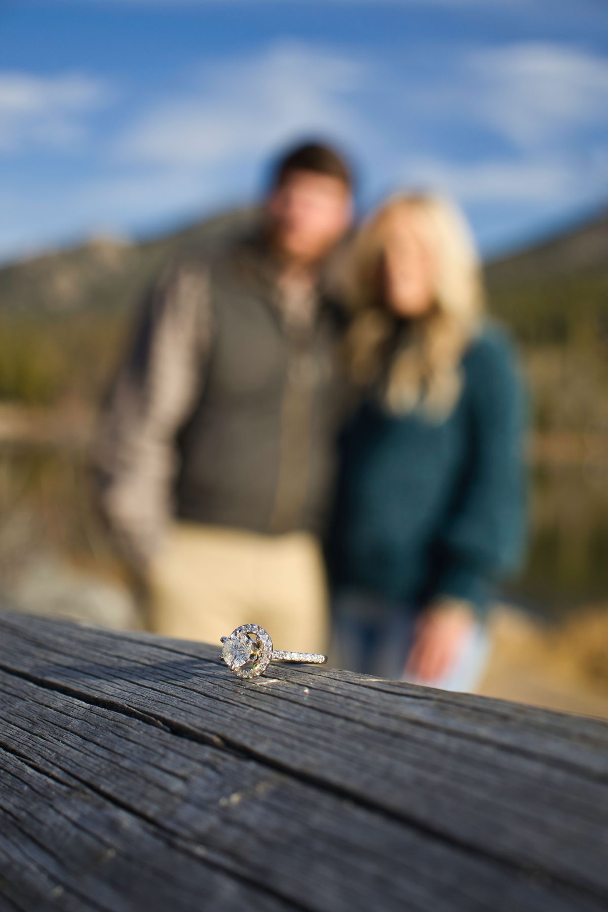 Engagement ring on the railing at Sprague Lake with the couple in the background. Pixcapades | The Rocky Mountain Proposal Photographer