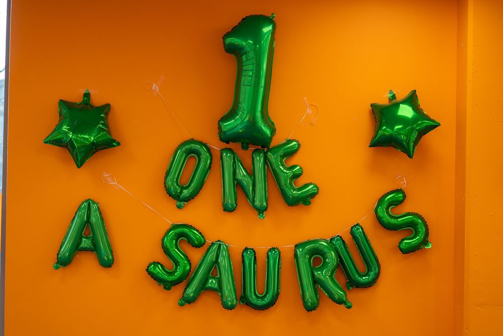 Green metallic balloon letters spelling 'ONE A SAURUS' with star balloons against an orange wall for dinosaur themed party.