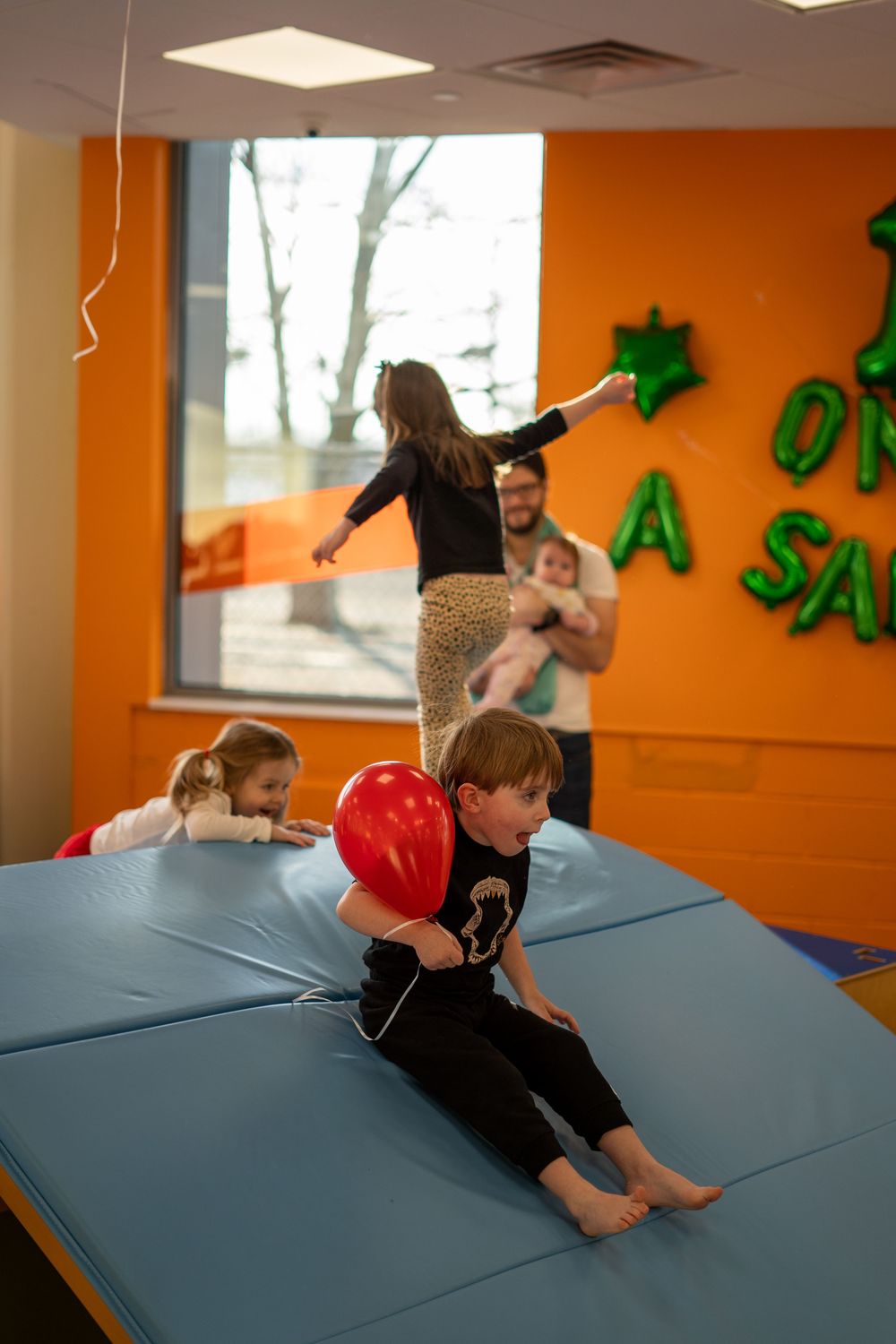 A child in black performs a sequence of forward rolls on a blue gymnastics mat in an orange-walled room.