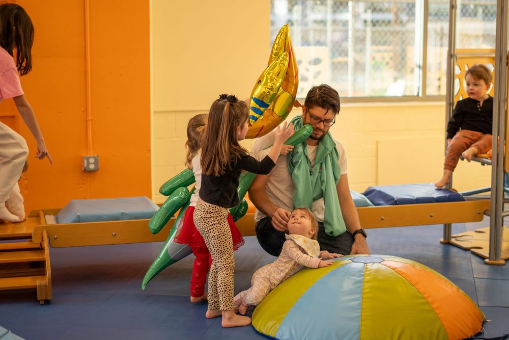 Children participate in an active indoor gymnastics class on colorful mats in a bright orange-walled room.