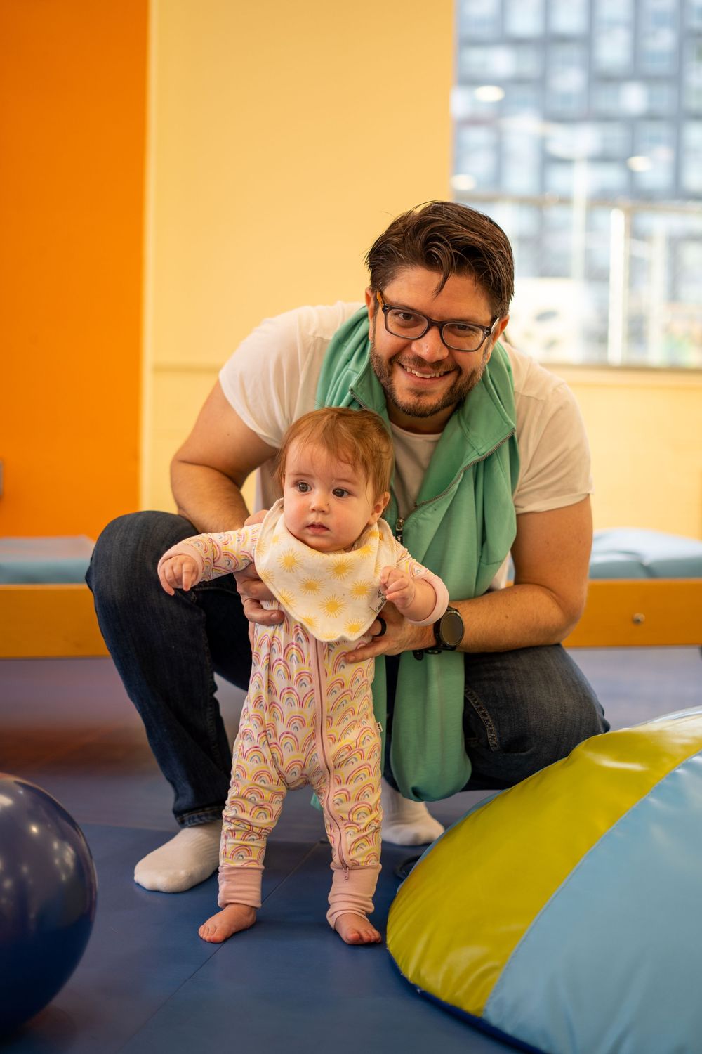 A caregiver helps support a baby learning to crawl and balance on colorful foam mats in an indoor play area.