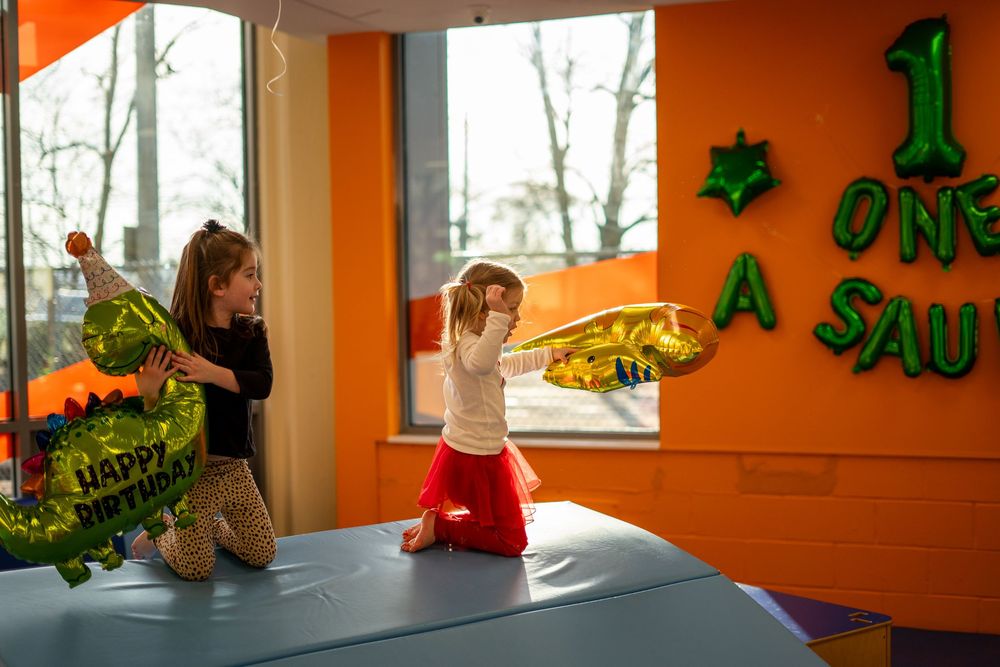 Children play with dinosaur balloons in a bright orange room with large windows during what appears to be a birthday party.