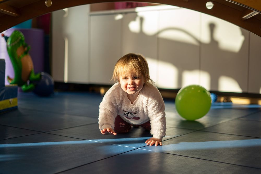 A toddler crawls on a blue mat in a sunlit room with toys scattered around.