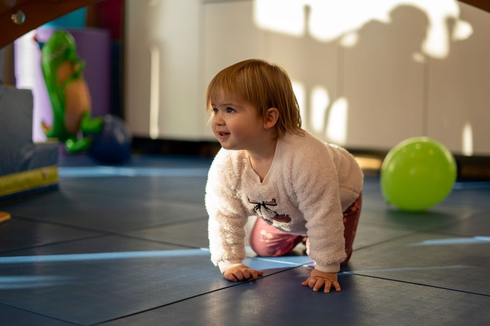 A toddler in a cream-colored sweater crawls on a dark floor mat with a green ball visible in the background.