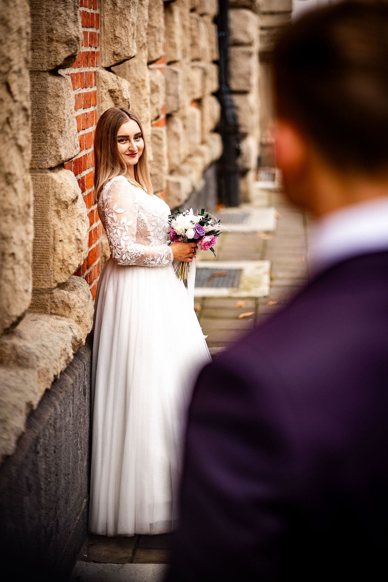 Bride in white lace dress poses against rustic stone wall.
