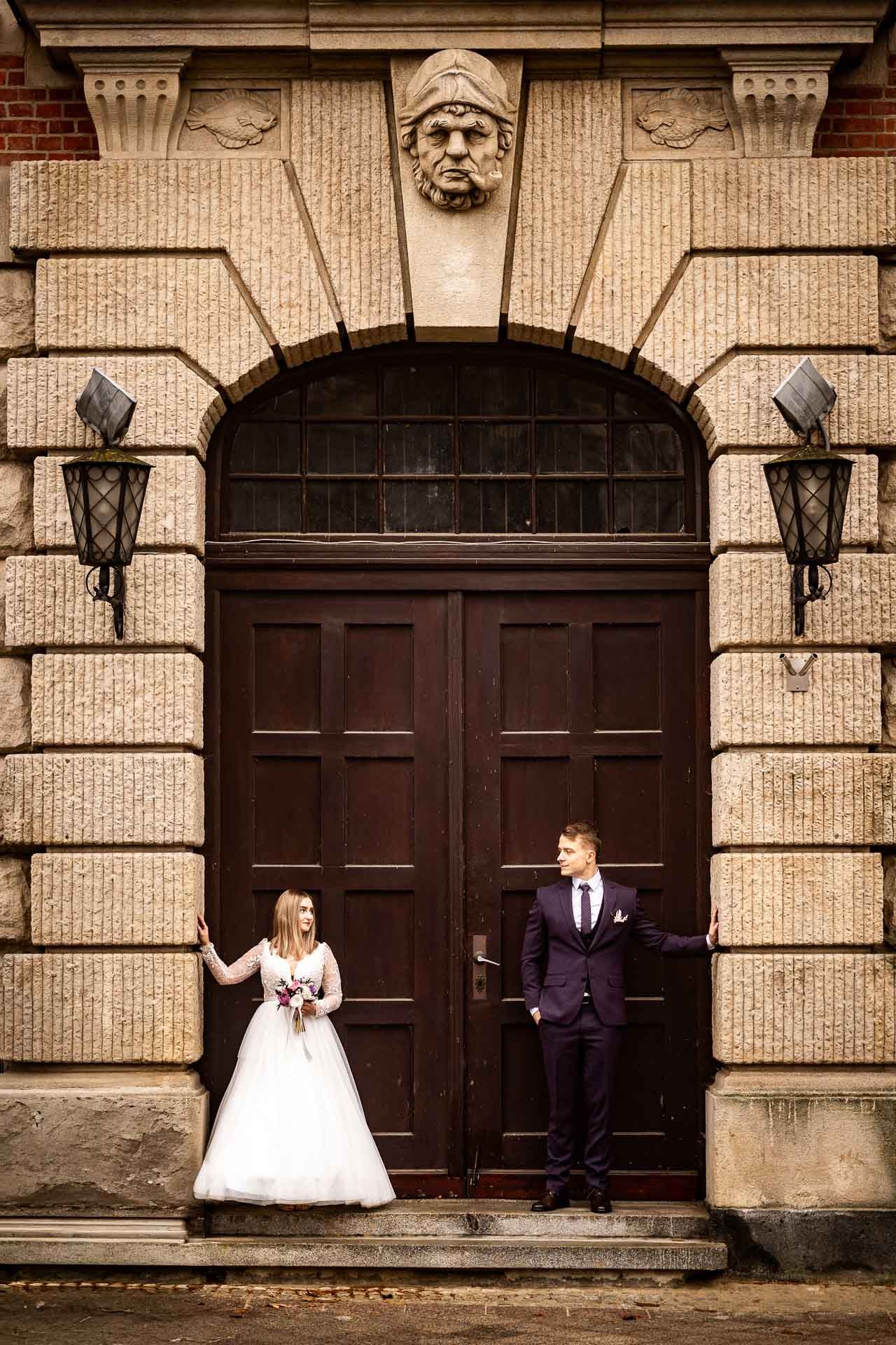 Wedding couple poses creatively against historic building with dark wooden doors.