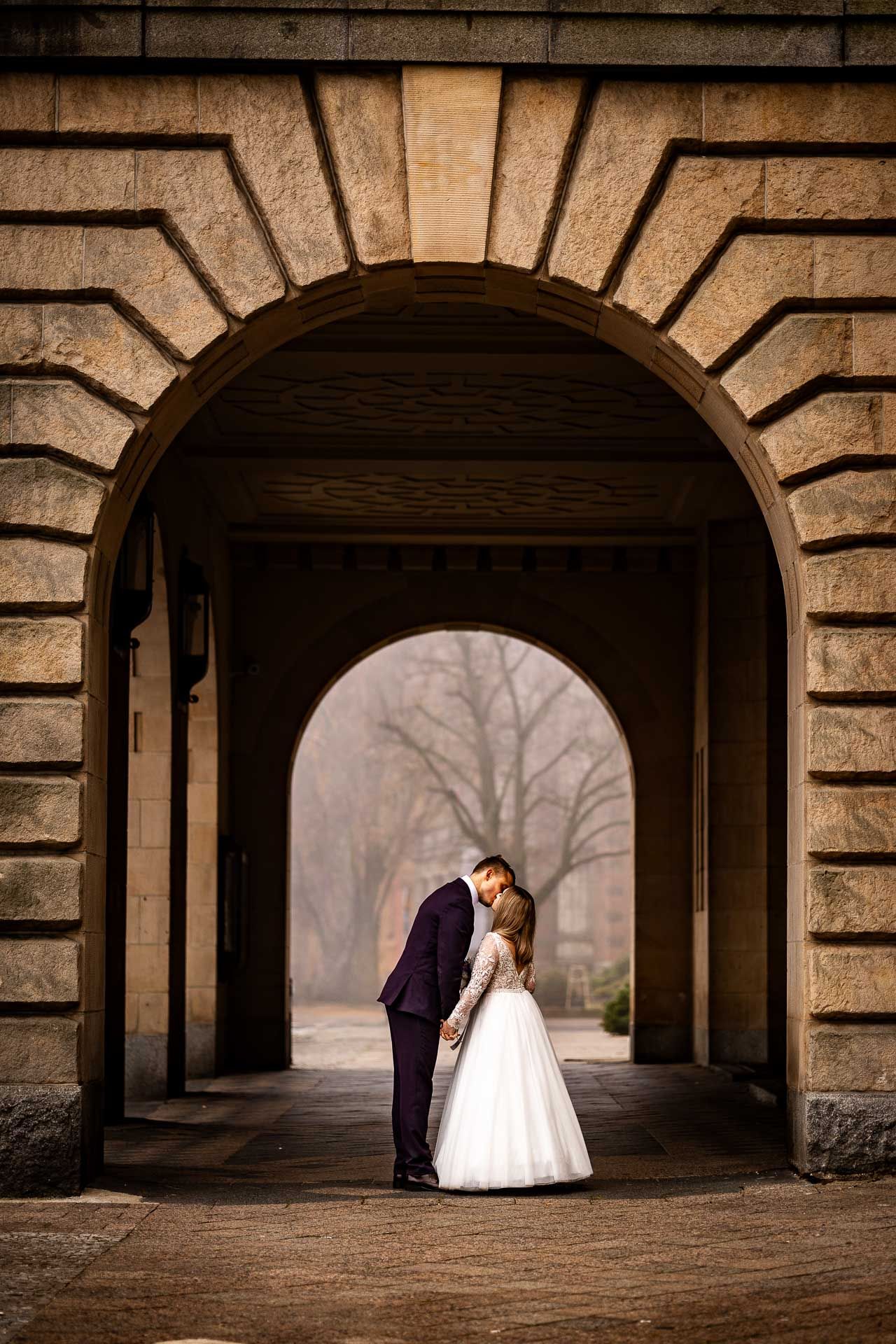 A romantic wedding couple embraces under a stone archway on a misty day.