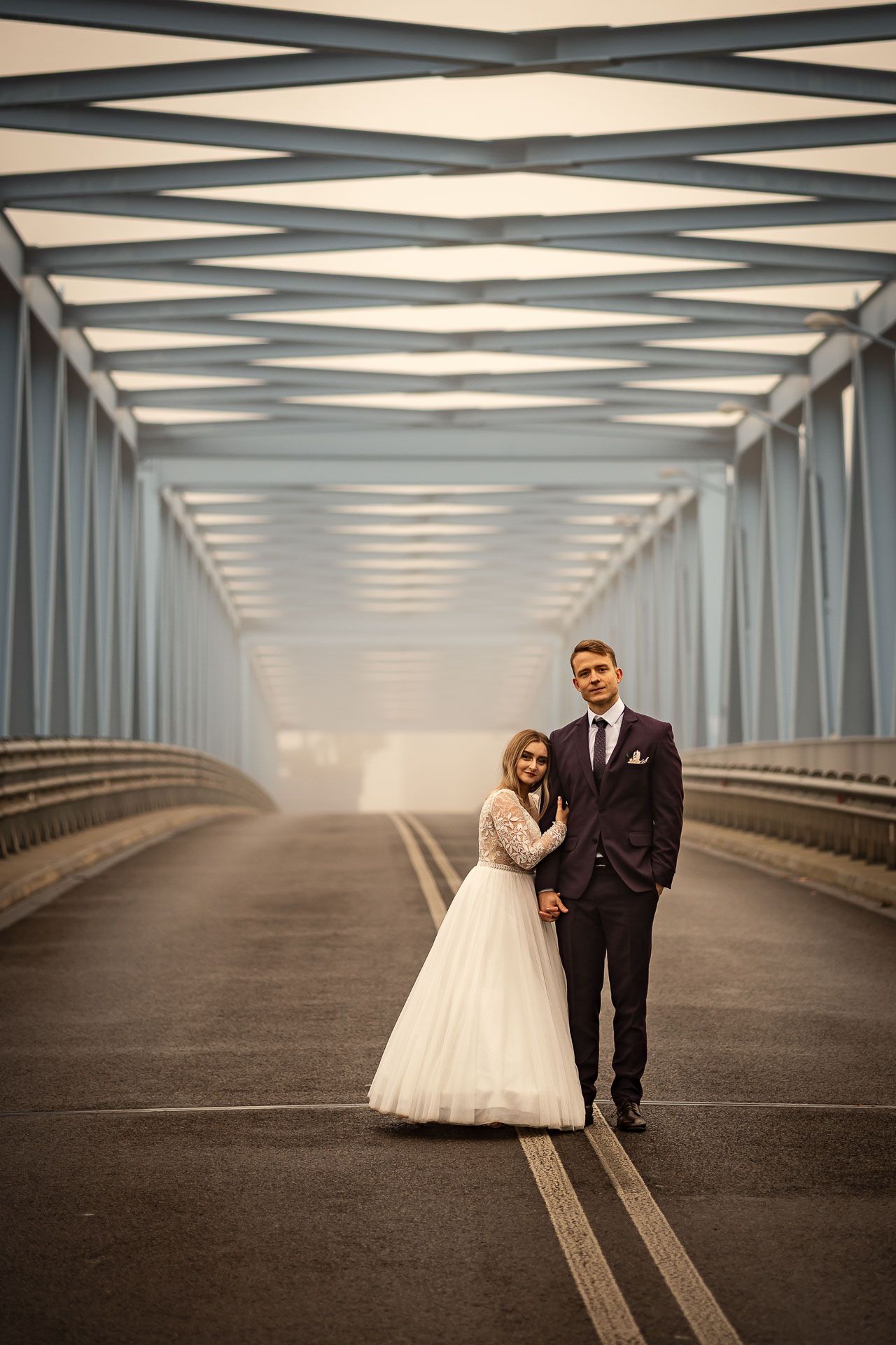 A bride and groom pose together on a modern bridge with repeating steel beams overhead.