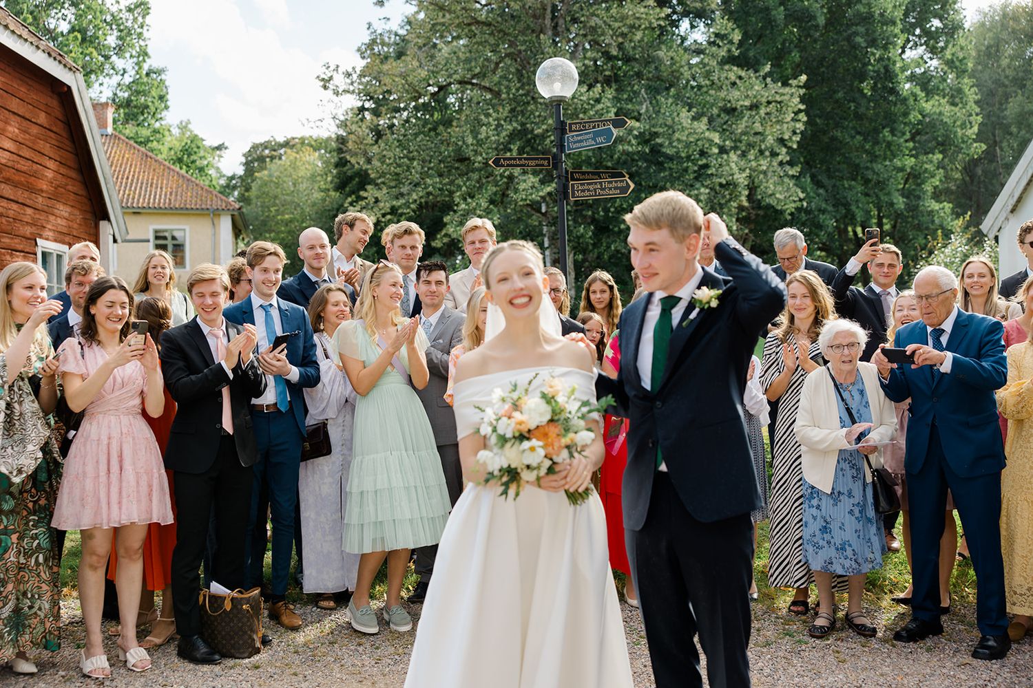 A wedding party and guests gather outdoors in front of a rustic wooden building during a summer celebration.