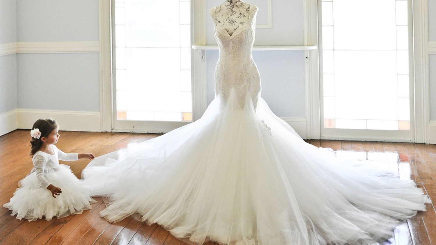 A young flower girl in white dress admires an elegant mermaid wedding gown displayed in a bright room.