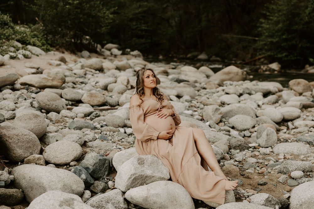 A person in a flowing peach dress poses elegantly on rocky riverbank in a scenic forest setting.