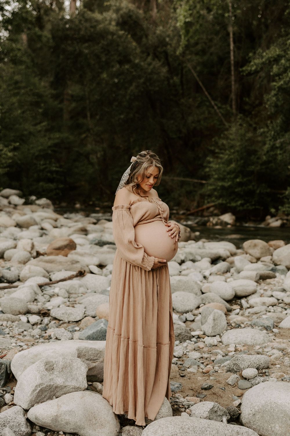 A pregnant woman in a flowing beige dress stands gracefully among large river rocks in a wooded setting.