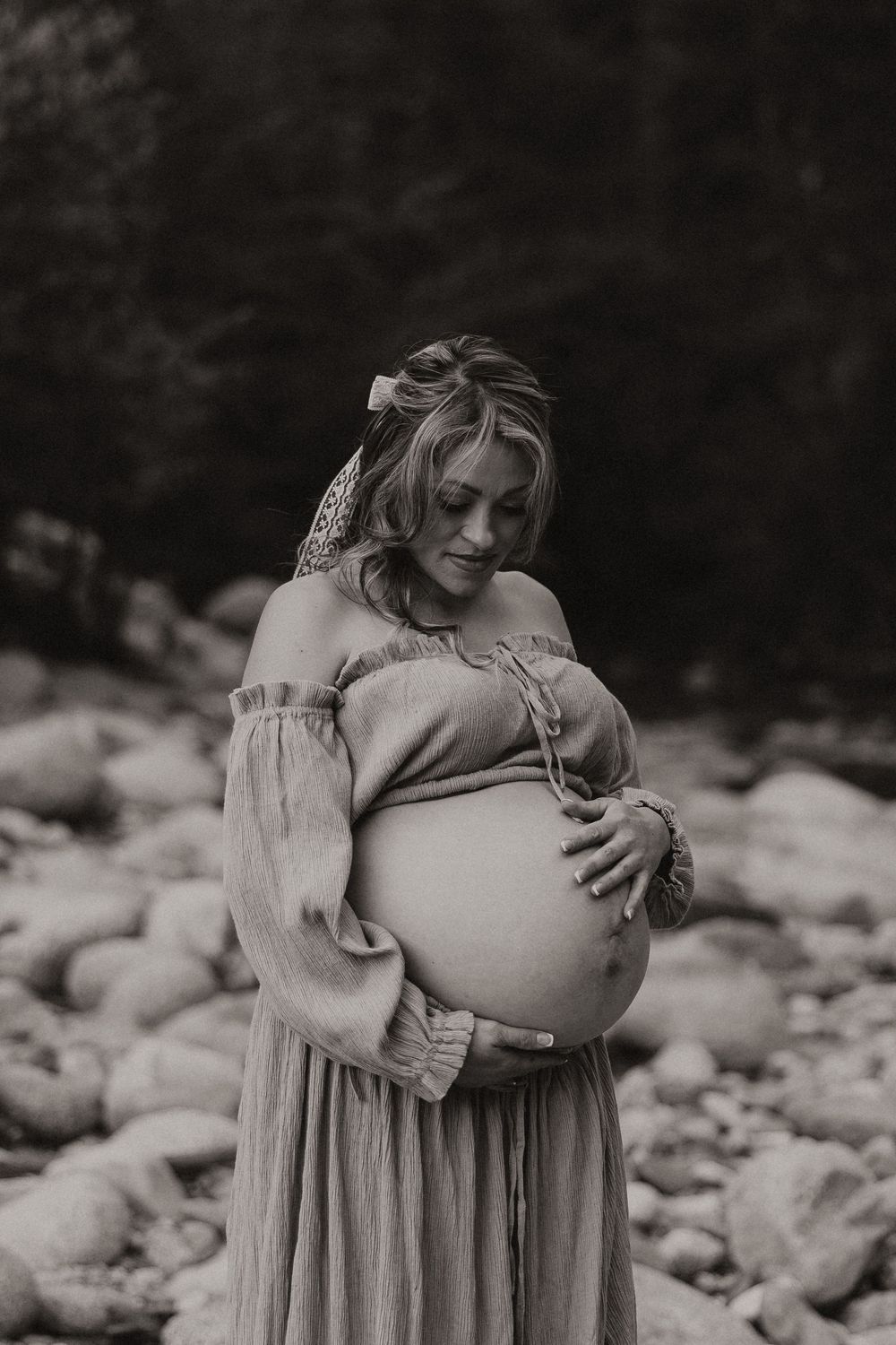 A pregnant woman in a flowy dress holds her belly while standing near a rocky riverbed in black and white.