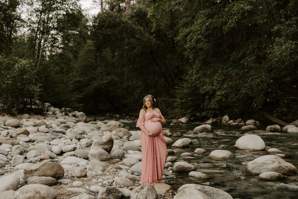 A romantic couple poses together in a natural setting with trees and rocks along a river.