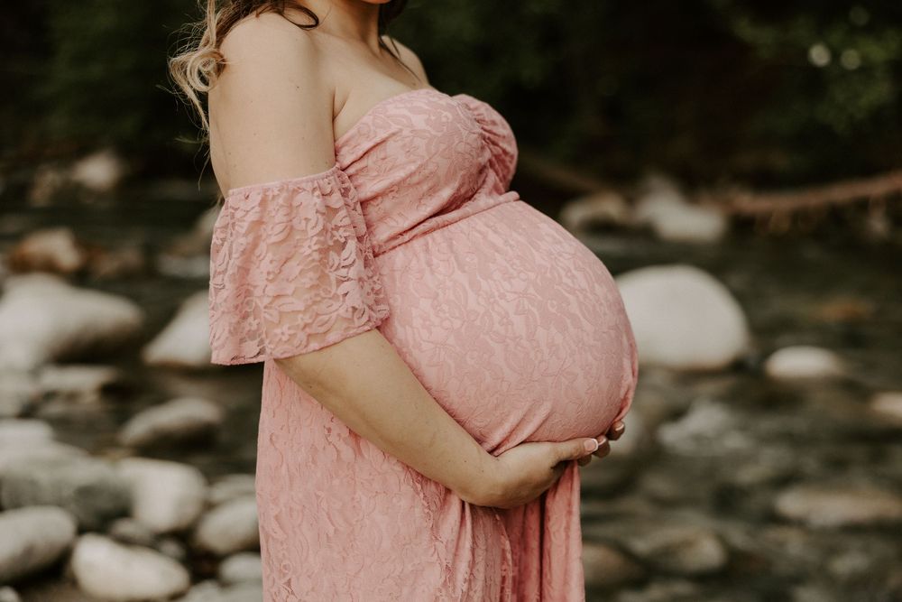 A person in a flowing pink lace maternity dress poses in a natural outdoor setting with rocks and greenery.
