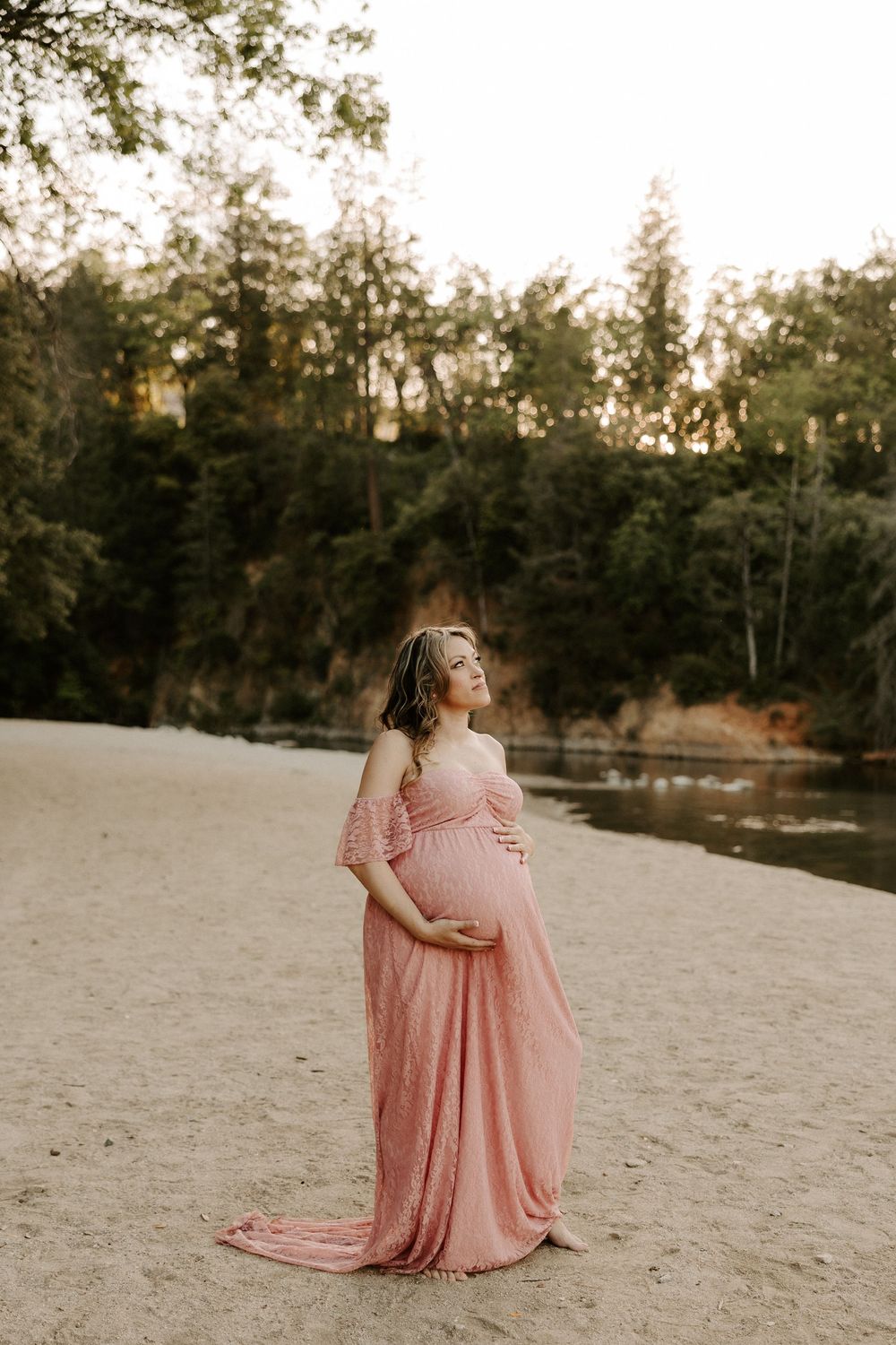 A person in a flowing pink dress sits gracefully on sandy ground with trees in the background during golden hour.