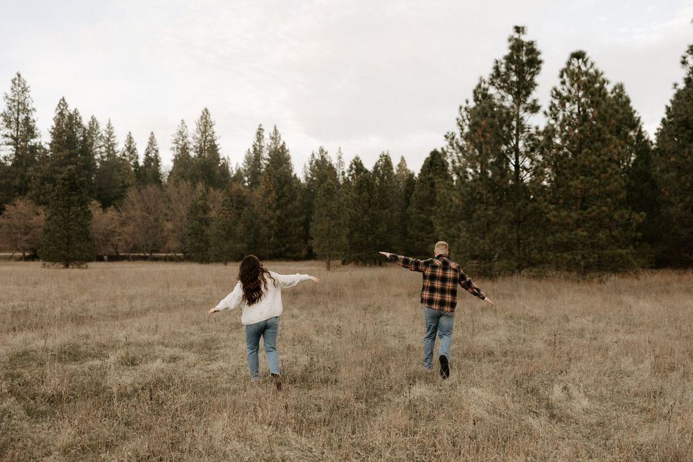 Two people walk through a golden field with evergreen trees in the background during autumn.