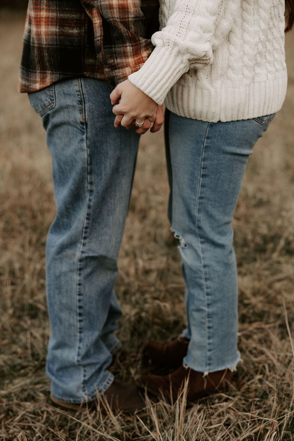 Close-up view of two people wearing blue jeans and cream sweaters standing together on dry grass in autumn.