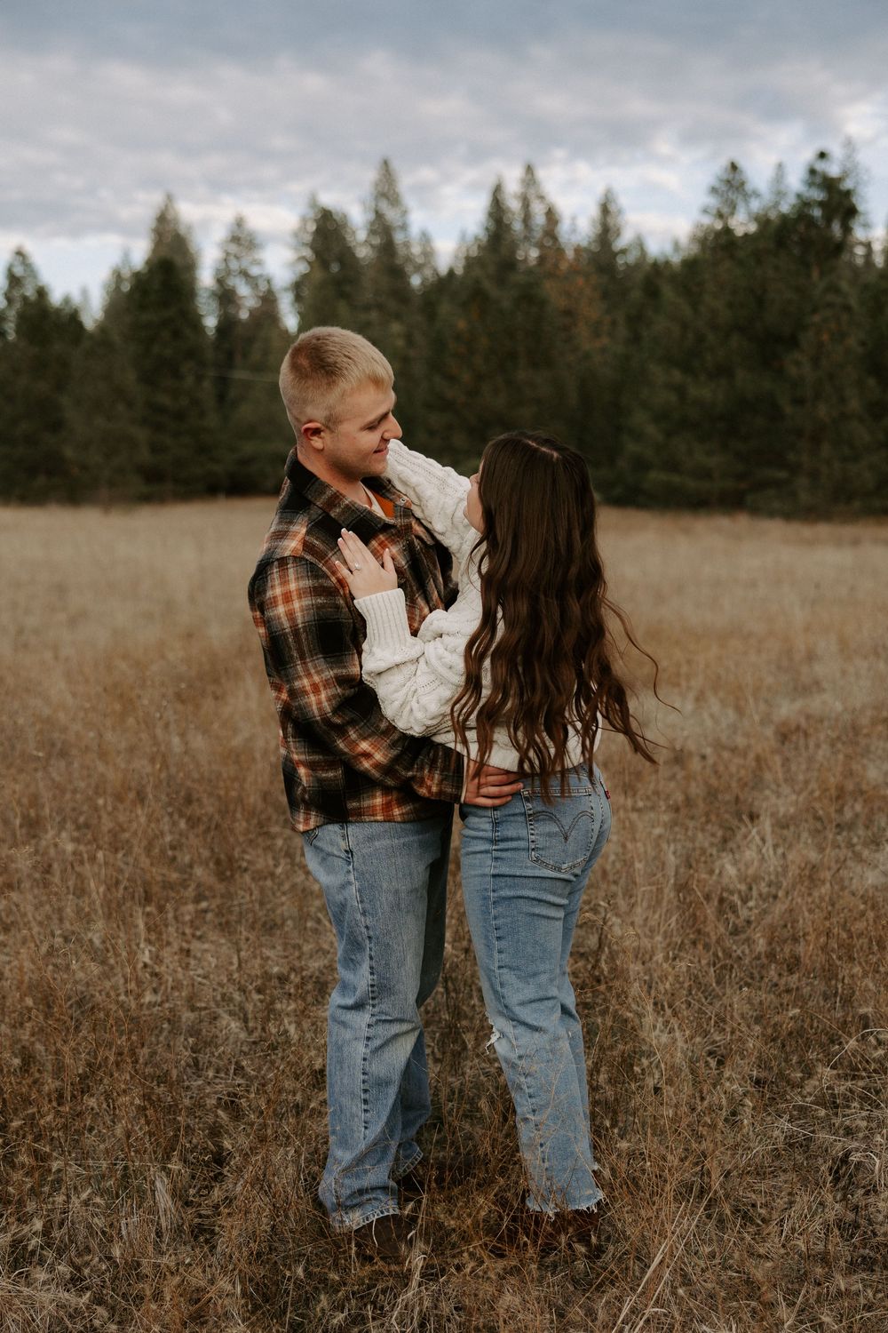 A couple in casual fall attire stands together in a golden field with evergreen trees in the background.