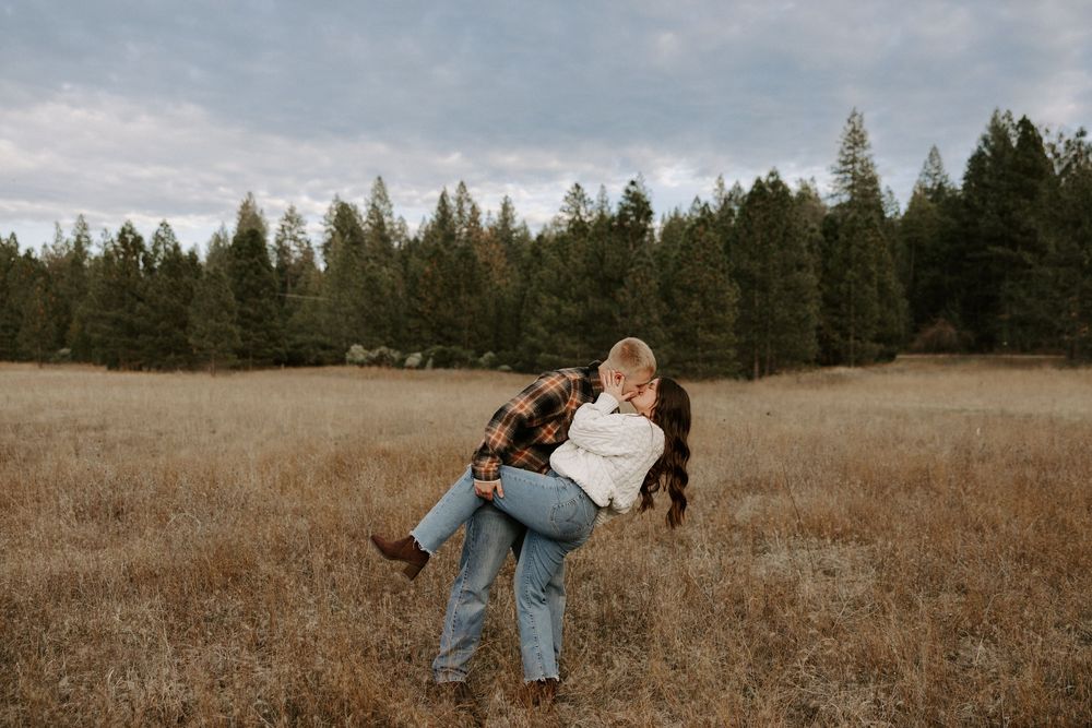A couple embraces in a rustic autumn field with tall grass and evergreen trees in the background.